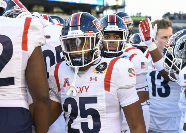 Gruppe von Männern in Fußballausrüstung auf einem Feld mit Helmen und Handschuhen, mit einer unscharfen Menge, Anzeigetafel und Himmel im Hintergrund.