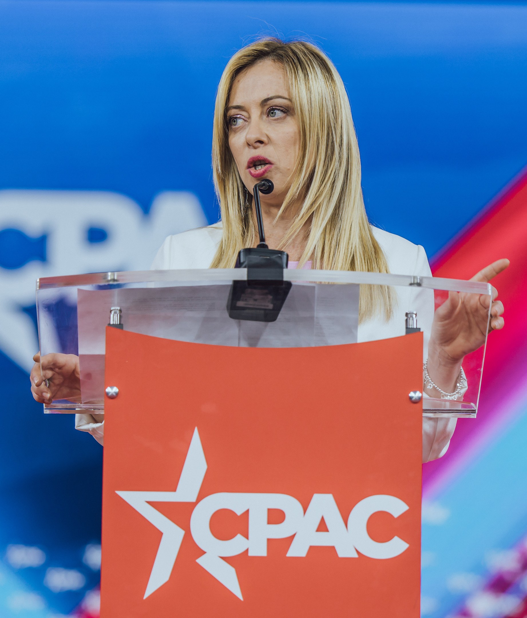 Frau mit Mikrofon an einem Rednerpult stehend, in einem weißen Kleid, als politische Wissenschaftlerin mit unscharfem Hintergrund.