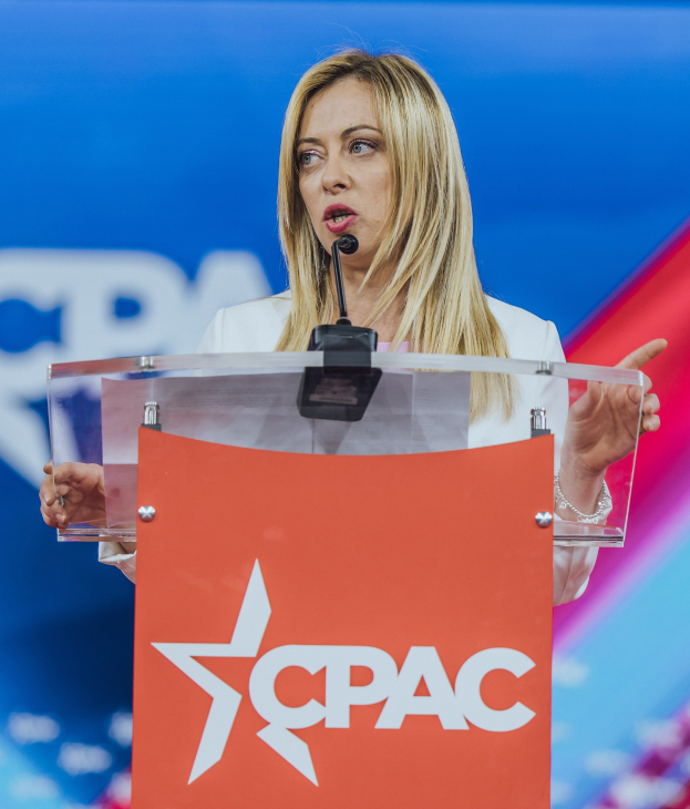 Frau mit Mikrofon an einem Rednerpult stehend, in einem weißen Kleid, als politische Wissenschaftlerin mit unscharfem Hintergrund.