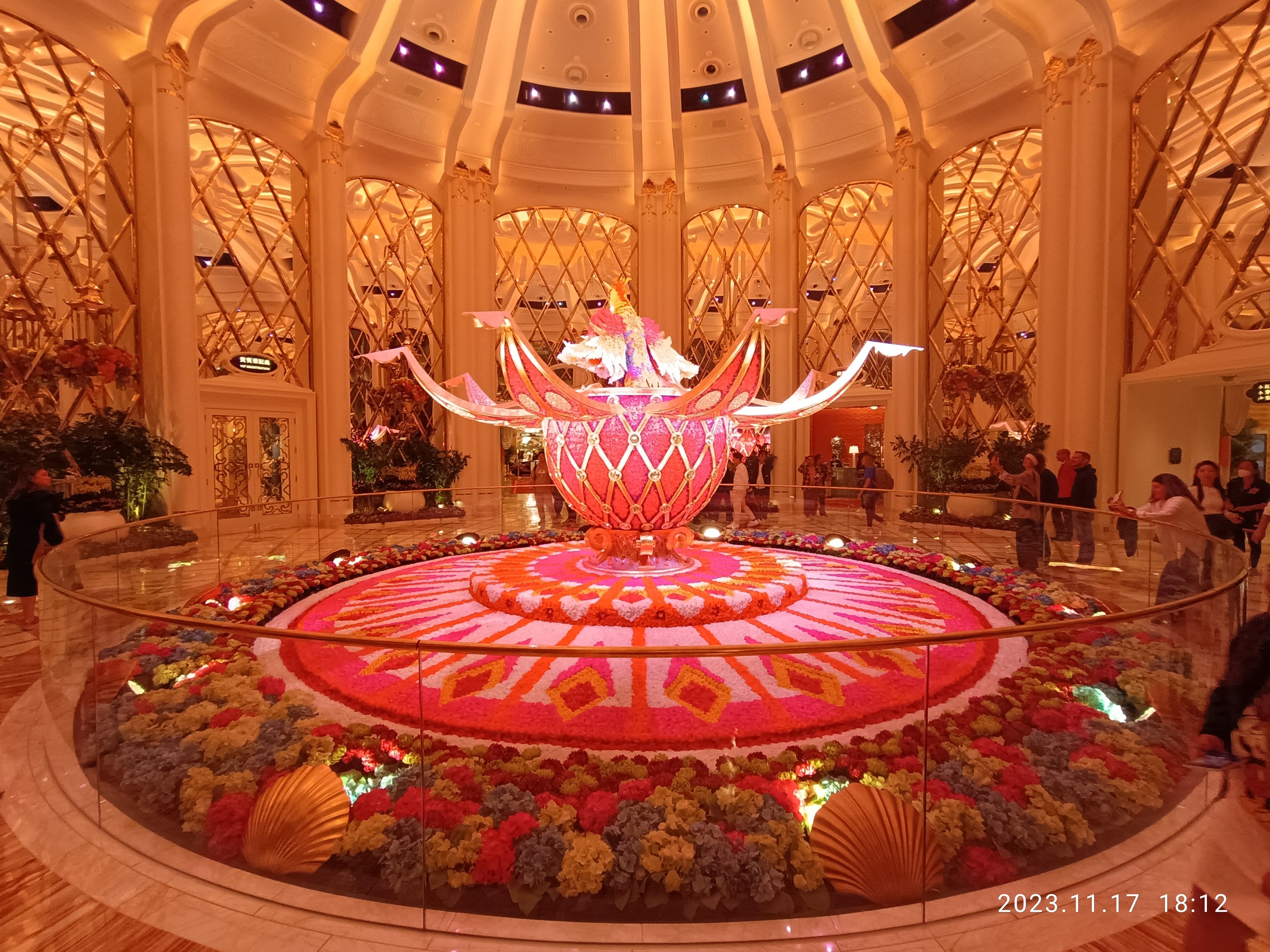 Exterior view of the Bellagio Hotel and Casino in Las Vegas, Nevada, featuring decorative floral arrangements on the floor, people with bags, and illuminated pillars in the background.