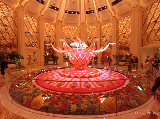Exterior view of the Bellagio Hotel and Casino in Las Vegas, Nevada, featuring decorative floral arrangements on the floor, people with bags, and illuminated pillars in the background.