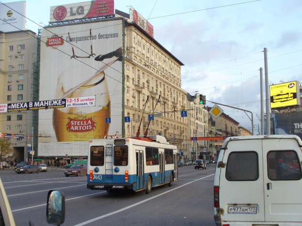 Eine belebte Stadtstraße mit verschiedenen Fahrzeugen, darunter ein Bus und ein Lieferwagen, Gebäuden mit Fenstern, Schildern, Strommasten mit Drähten und Verkehrsampeln.