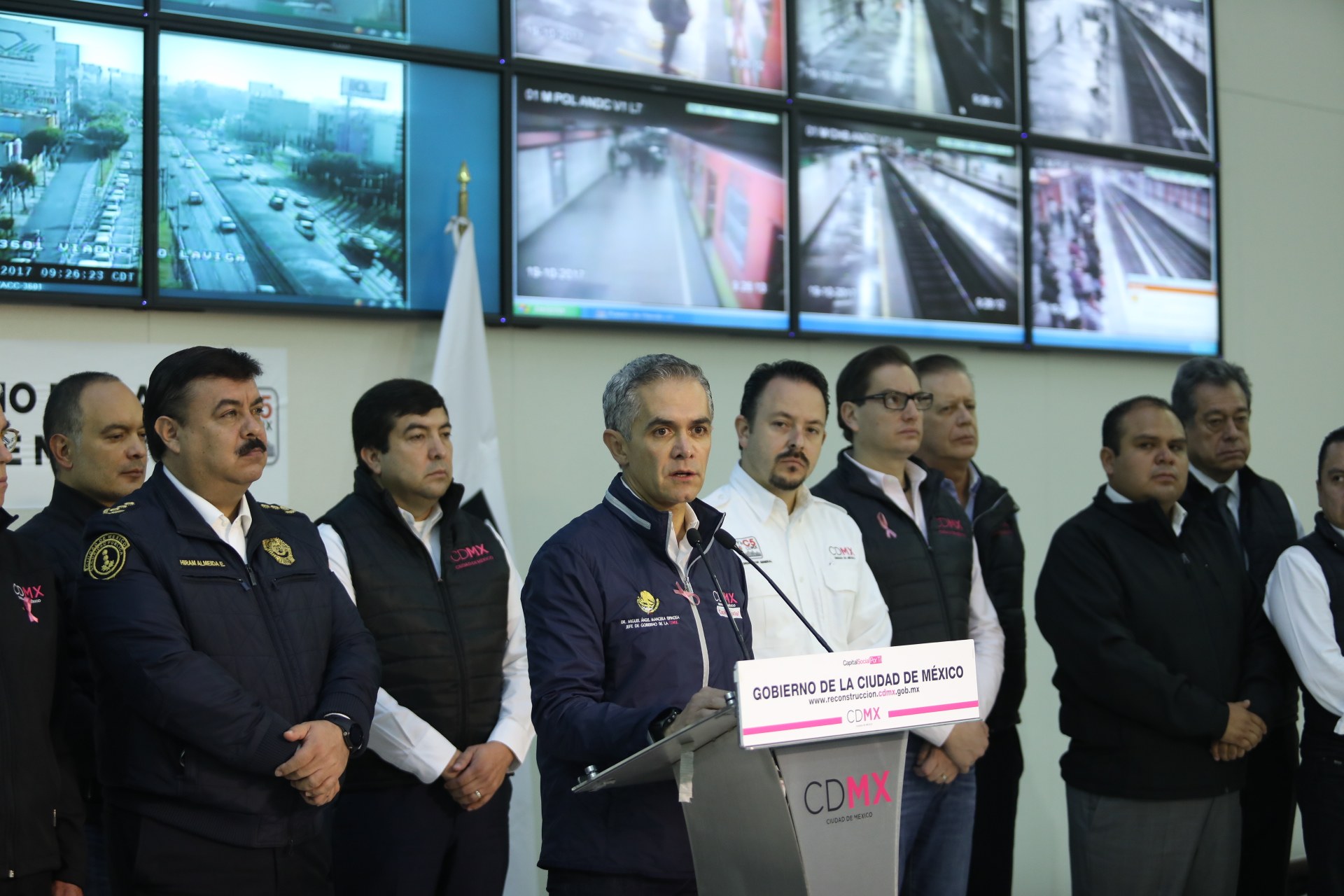 Gruppe von Männern, die vor einem Podium mit einem Mikrofon und einer Tafel mit der Aufschrift "Mexikanisches Zoll- und Grenzschutz" stehen, mit Fernsehgeräten und einer Flagge an der Wand dahinter.