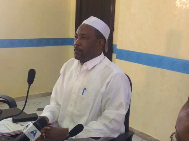 Ein Mann in einem weißen Kleid und Hut sitzt an einem Tisch mit Mikrofonen, spricht, während Papiere und Gegenstände auf dem Tisch liegen, mit einer anderen Person mit Brille rechts von ihm, in einem Raum mit einer Wand und einer Tür.