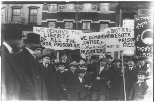 Schwarz-weißes Foto einer Gruppe von Menschen in einer Reihe mit Schildern, die "Wir fordern die Freiheit aller Gerechtigkeit für die Gefangenen" tragen, vor einem Gebäude mit Fenstern und einem Geländer.
