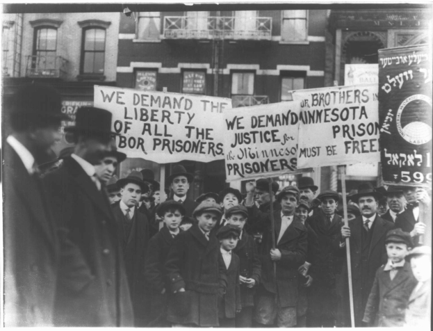 Schwarz-weißes Foto einer Gruppe von Menschen in einer Reihe mit Schildern, die "Wir fordern die Freiheit aller Gerechtigkeit für die Gefangenen" tragen, vor einem Gebäude mit Fenstern und einem Geländer.