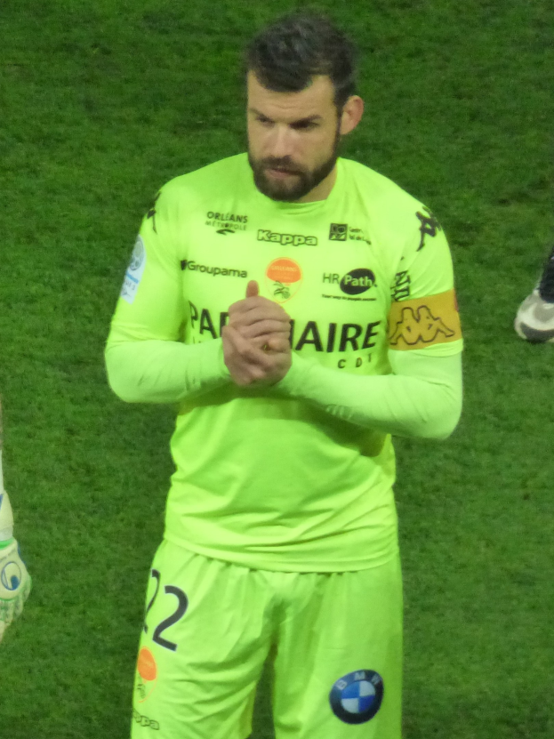 Ein Torwart in einem gelben Trikot klatscht auf einem Fussballfeld, trägt ein grünes Hemd und Shorts und hat ein weiteres Bein links sichtbar.