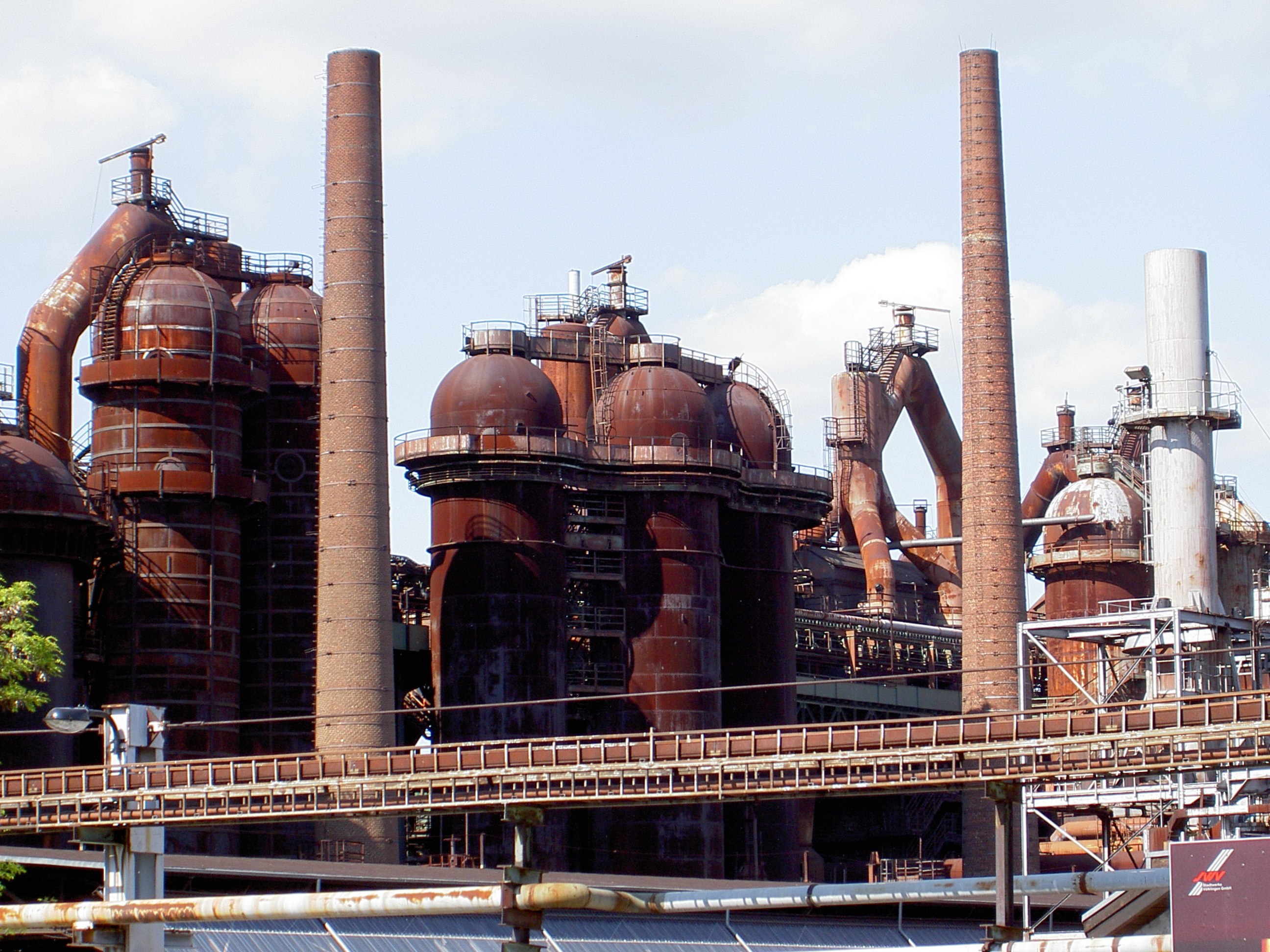 Großes Industriegelände mit zahlreichen Rohren, Schornsteinen, einer Brücke, Strommasten, Straßenlaternen, umliegenden Bäumen und einem bewölkten Himmel.