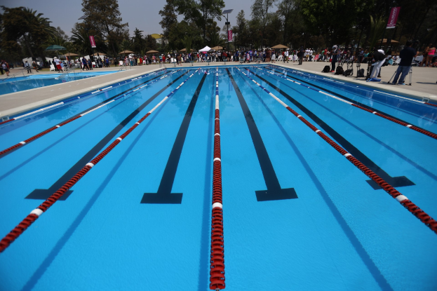 Großer Schwimmbad mit Bahnentrennungen, umgeben von Menschen mit Taschen und Gegenständen, mit Zelten, Schildern, Masten, einem Schild und einer bewölkten Himmel im Hintergrund.