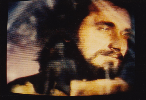 Ein Mann mit dunklem Bart und Schnurrbart, langen dunklen Haaren und einem ernsten Gesichtsausdruck in einem weißen Hemd ist auf einem Fernsehbildschirm mit dunklem Hintergrund zu sehen.