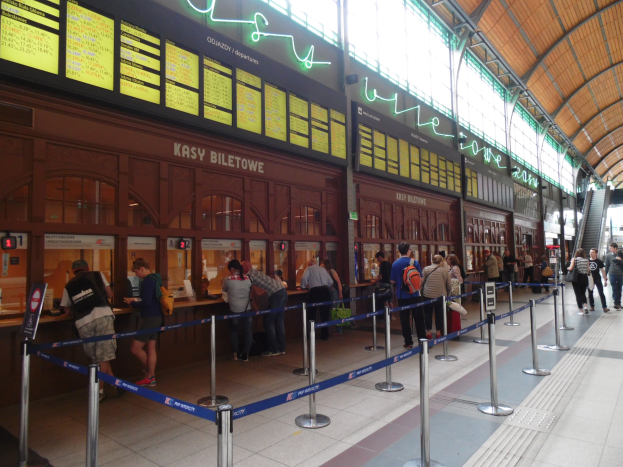 Menschen in einer Warteschlange an einem Bahnhofsschalter mit Pfosten, Bändern, Informationsschildern, Deckenleuchten und Treppen mit Geländern.