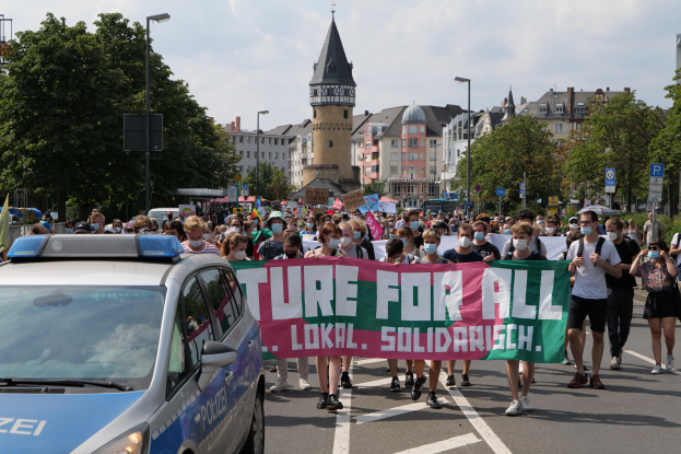 Eine große Gruppe von Menschen marschiert mit einem Banner mit der Aufschrift "Lokale Solidarität" durch eine Straße, begleitet von einem Polizeiwagen, Bäumen, Laternen, Schildern, Gebäuden und einem bewölkten Himmel.