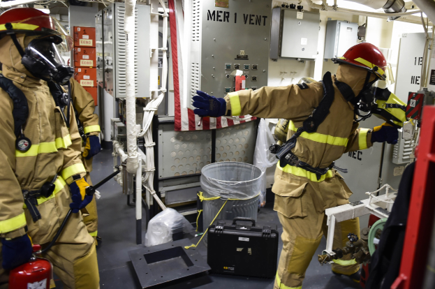 Drei Feuerwehrleute in Schutzausrüstung stehen zusammen in einem Raum mit einem Feuerlöscher links, einer Maschine rechts und diversen Gegenständen auf dem Boden.