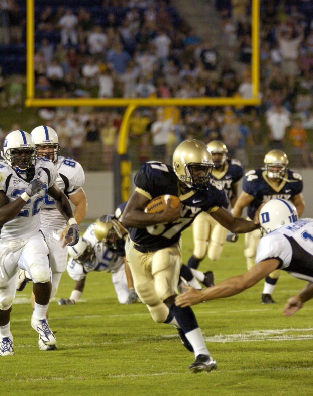 Ein Fußballspieler in Uniform rennt mit dem Ball, umgeben von Spielern in Sportbekleidung und Helmen während eines Spiels.