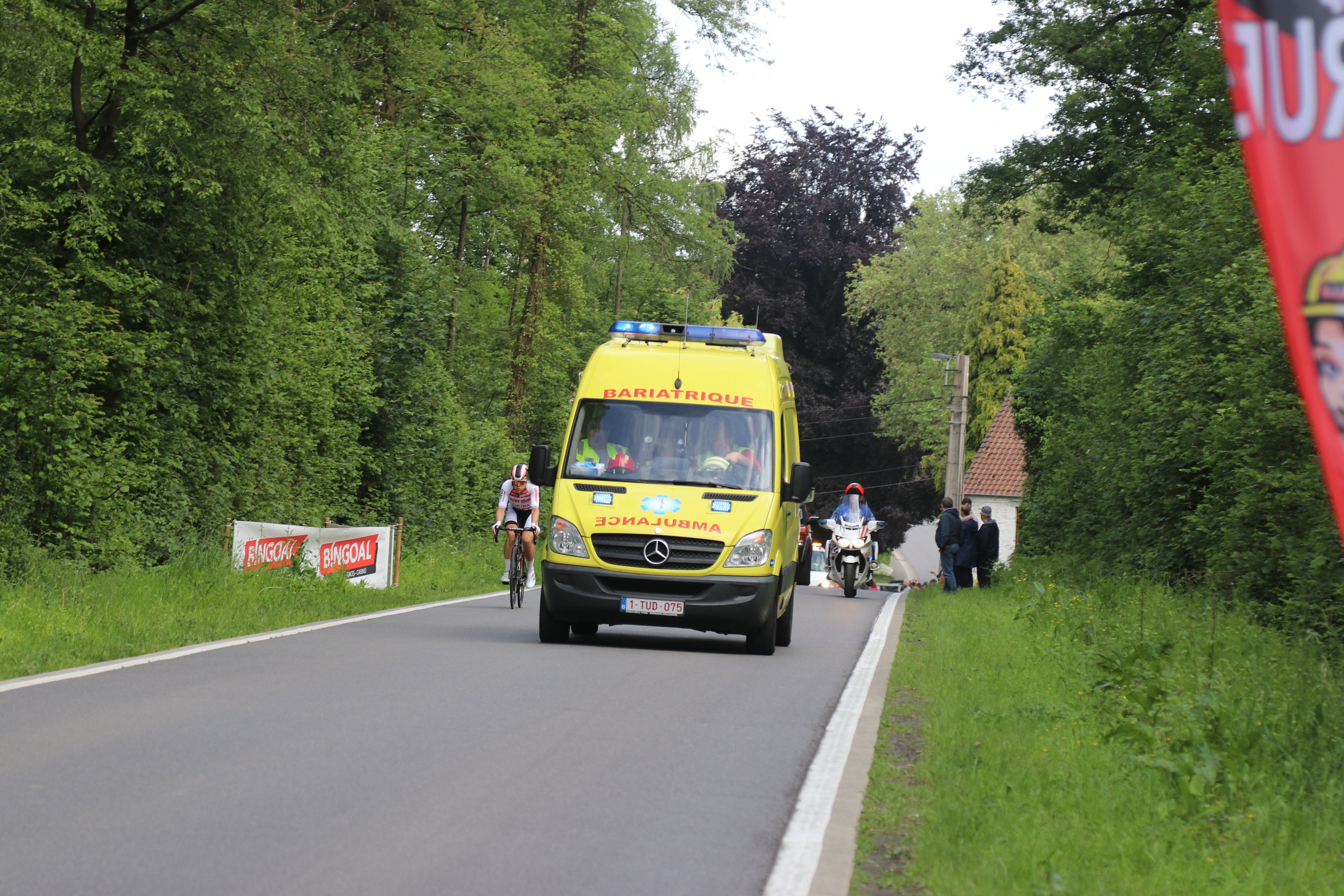 Ambulanz fährt auf einer Straße mit Radfahrern daneben, umgeben von Gras, Bäumen, Häusern, Strommasten und einem klaren blauen Himmel.