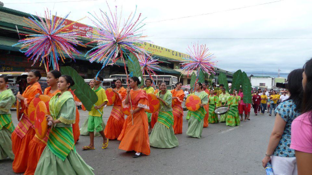 Eine Gruppe von Menschen in bunten Kostümen marschiert während eines Karnevalsumzugs durch die Straße, wobei eine Frau auf der rechten Seite eine Flasche hält, und Fahrzeuge, Gebäude, Bäume, Pfähle, Drähte und der Himmel im Hintergrund zu sehen sind.