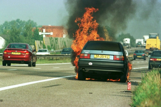 Ein Auto in Flammen am Straßenrand mit anderen Fahrzeugen in der Nähe, Bäumen, Gebäuden und einem klaren blauen Himmel im Hintergrund und einem Feuerlöscher rechts.
