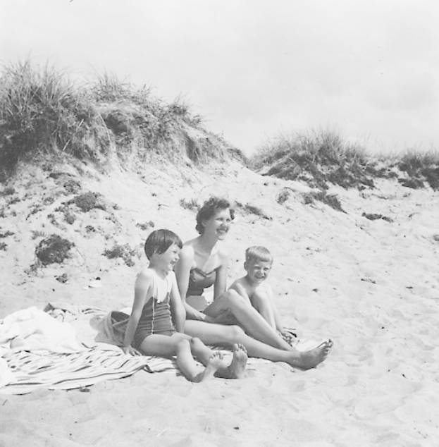 Schwarz-weiß-Foto einer Frau in einem Bikini und zwei Kindern in Badebekleidung auf einer Decke am Strand mit Sand, Gras und Himmel im Hintergrund.
