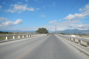 Eine Autobahn mit Motorradfahrern darauf, umgeben von Geländern, Strommästen mit Käble, Bäumen, Hügeln und einem bewölkten Himmel im Hintergrund.