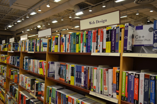 Ein Bibliotheksinneres mit Reihen von Bücherregalen, Informationsschildern, Deckenbeleuchtung und sichtbaren Rohren an der Decke.