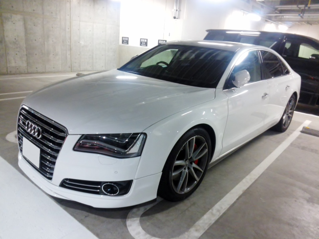 Ein weißer Audi S8, der in einer Tiefgarage neben einem schwarzen Auto geparkt ist, mit sichtbaren Garagenwänden, Deckenrohren und Leuchten im Hintergrund.