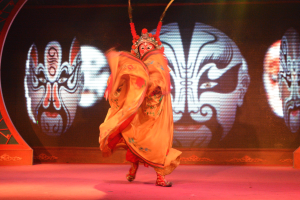 Eine Person in einem chinesischen Opernkostüm und einer Maske tritt auf der Bühne mit ausgestreckten Armen und zurückgelegtem Kopf auf, während ein Bildschirm im Hintergrund Bilder anzeigt.