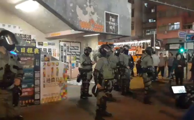 Polizisten in Schutzausrüstung stehen vor einer Menge von Helmet tragenden Demonstranten mit Waffen, mit Plakaten an Wänden, Deckenleuchten und Gebäuden im Hintergrund in Hong Kong.