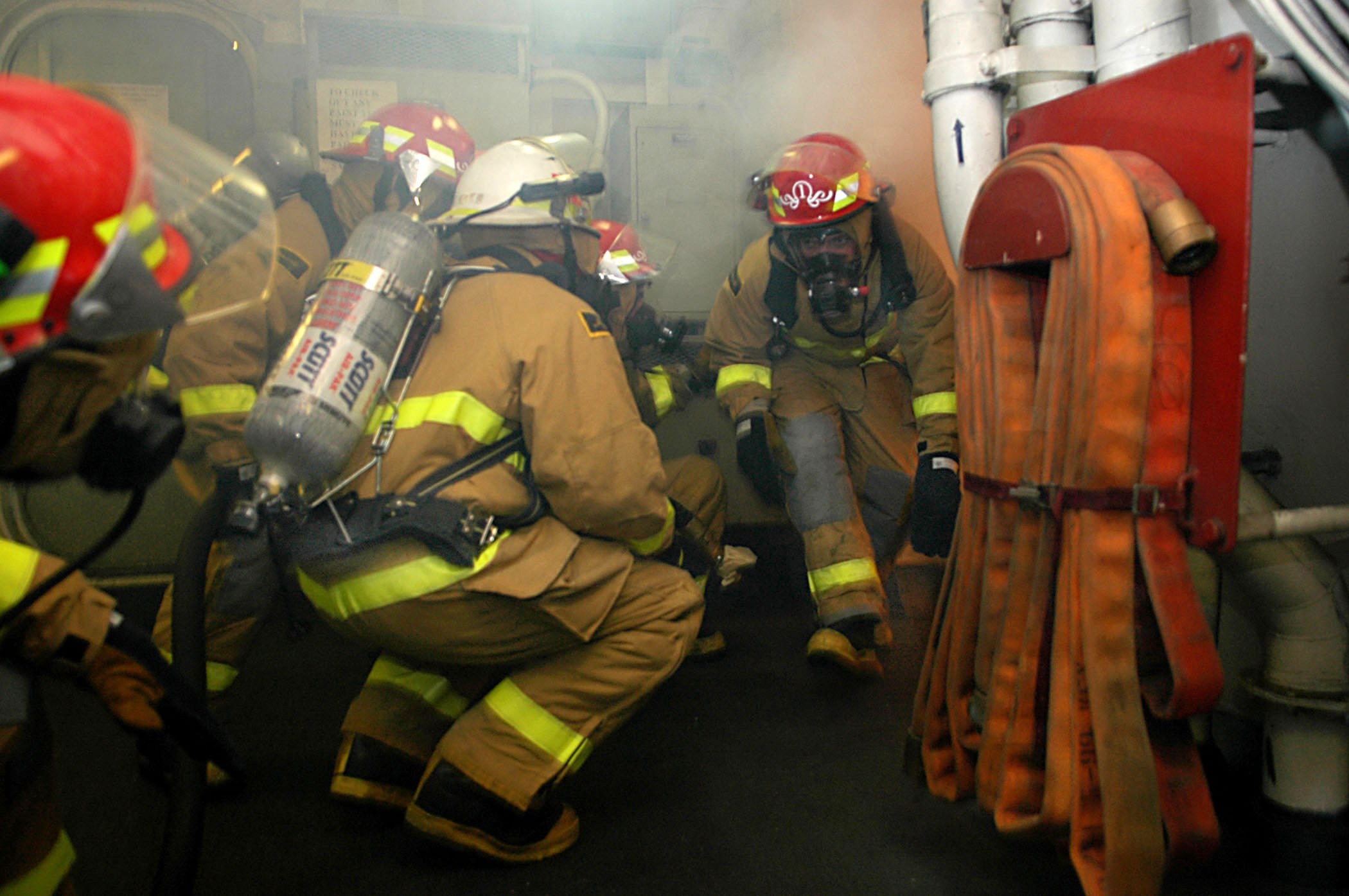 Feuerwehrleute in Schutzausrüstung mit Rauch aus dem Mund, die in einem Raum mit Rohren und einer Wand im Hintergrund stehen.