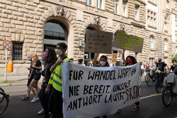 Eine Gruppe von Menschen nimmt an einer Protestdemo teil und marschiert durch eine Straße, einige halten Schilder und andere fahren Fahrräder, im Hintergrund ist ein Gebäude mit Fenstern, Bögen, Säulen und Skulpturen zu sehen.