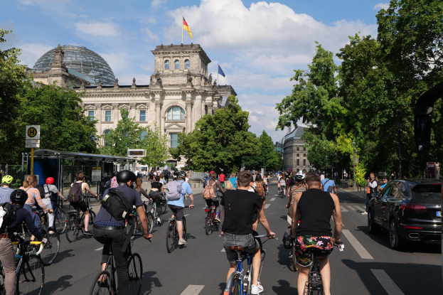 Eine Gruppe von Menschen, die auf Fahrrädern eine von Bäumen gesäumte Straße in Berlin entlangfahren, mit Gebäuden und einer Bushaltestelle im Hintergrund, unter einem bewölkten Himmel und einer Flagge an einem Gebäude.