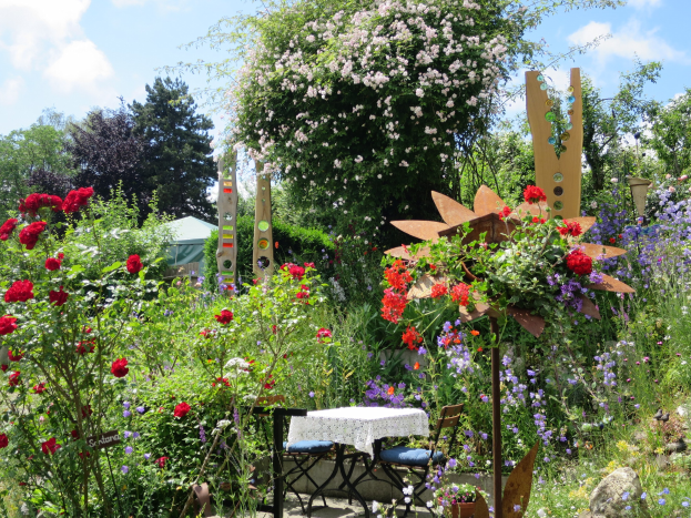 Ein üppiger Garten auf der Chelsea Flower Show mit einem zentralen Tisch mit einer Decke, umgeben von bunten Blumen und Pflanzen, mit Bäumen und einem bewölkten Himmel im Hintergrund.
