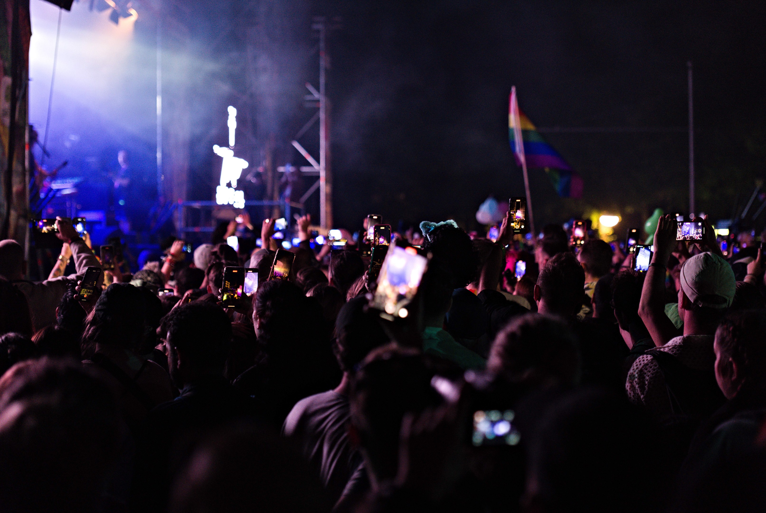 Eine Menge bei einem Konzert mit hochgehaltenen Handys, beleuchtet von Scheinwerfern, umgeben von Pfosten und einer Fahne, während Musiker auf der Bühne spielen.
