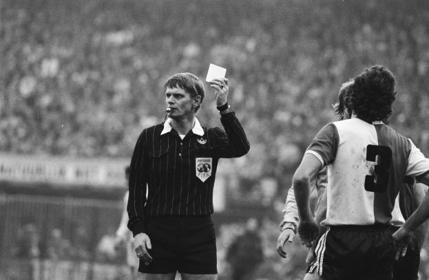 Schwarzes und weißes Bild eines Schiedsrichters, der eine rote Karte in einer Menge von Menschen mit Fußballuniformen hält, mit einem Zaun im Hintergrund.