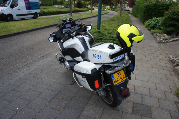 Ein Polizeimotorrad am Straßenrand mit einem Helm darauf, umgeben von Bäumen, Polen, Gebäuden, Pflanzen, Gras, Steinen und anderen Fahrzeugen im Hintergrund.