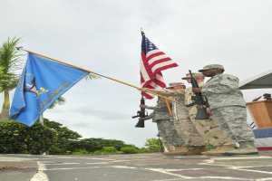 Eine Gruppe von Männern in Militäruniformen mit Gewehren, die neben einem Podium mit Mikrofon stehen, mit Bäumen und einem klaren blauen Himmel im Hintergrund.