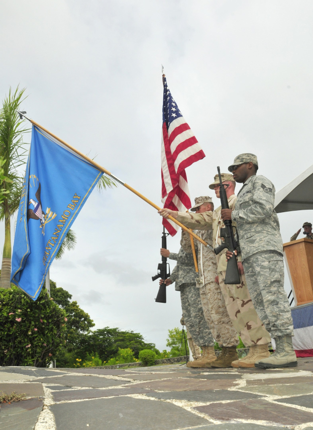 Eine Gruppe von Männern in Militäruniformen mit Gewehren, die neben einem Podium mit Mikrofon stehen, mit Bäumen und einem klaren blauen Himmel im Hintergrund.