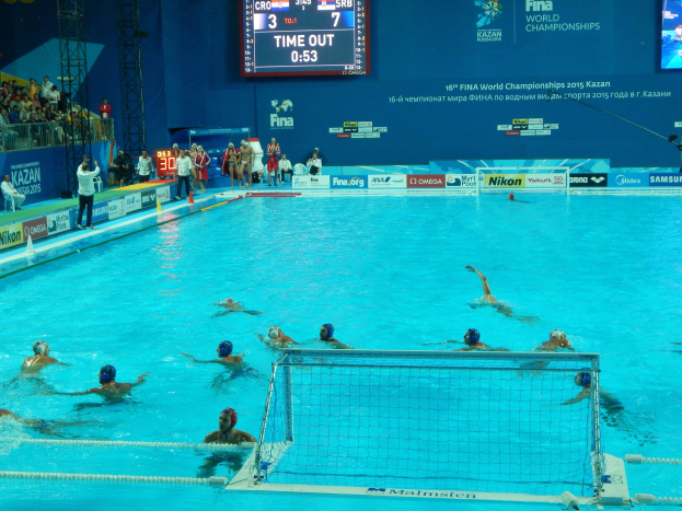 Gruppe von Athleten, die Wasserball im Pool während der FINA-Weltmeisterschaften 2015 in Kazan spielen, mit Zuschauern und Eventbeschilderung im Hintergrund.