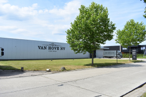 Eine Van Hool NV-Anlage in den Niederlanden mit Fahrzeugen, Gras, Bäumen, Gebäuden, Schildern, Pfählen und einem bewölkten Himmel im Hintergrund.