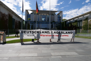 Eine Gruppe von Menschen, die ein Transparent mit der Aufschrift "Deutschland, Lagerland Migration ist kein Verbrechen" vor einem Fensterbau mit Fenstern hölt, umgeben von Gras, einem Metallzaun, Pflanzen, Bäumen und Fahnenständen unter einem bewölktem Himmel.