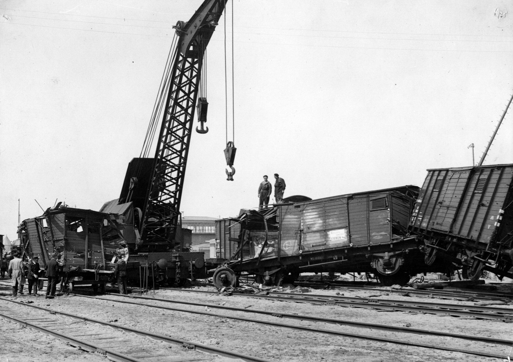 Schwarzes und weißes Bild eines Zugunglücks mit einem Kran, der einen Zugwagen auf die Schienen hebt, mehrere Personen in der Nähe und im Hintergrund Gebäude und einen klaren Himmel.