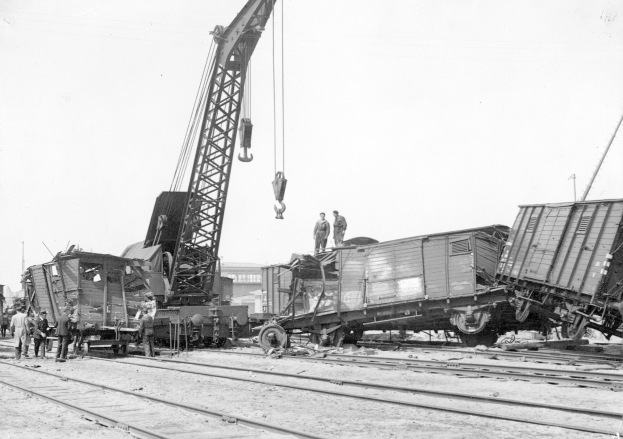 Schwarzes und weißes Bild eines Zugunglücks mit einem Kran, der einen Zugwagen auf die Schienen hebt, mehrere Personen in der Nähe und im Hintergrund Gebäude und einen klaren Himmel.