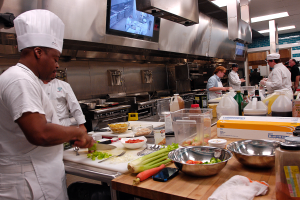 Eine Gruppe von Köchen in einer Küche, die Essen auf einem Schneidbrett zubereiten, mit einem Koch im Vordergrund in einem weißen Kleid und einer Mütze, der ein Messer hält und von Utensilien, Schüsseln, Gemüse, einem Handy, Flaschen und anderen Gegenständen umgeben ist, während im Hintergrund ein Fernseher und Deckenlampen zu sehen sind.