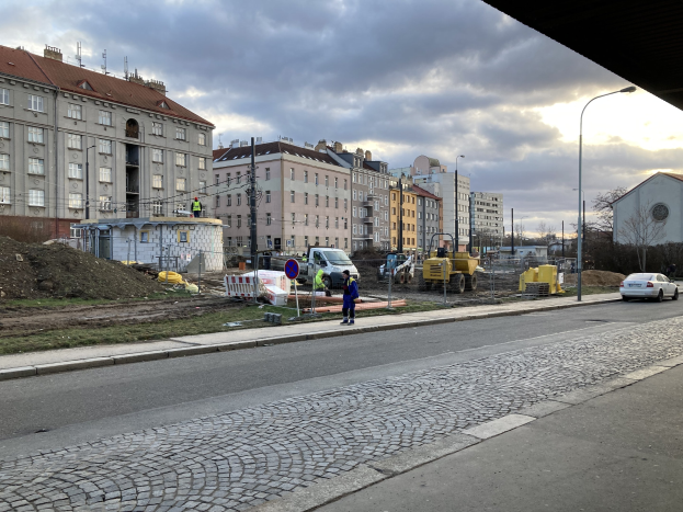 Eine Straße in einer Stadt mit parkenden Autos, Gebäuden, Laternen, Bäumen und einem bewölktem Himmel, mit einer Absperrung mit Fahrzeugen und Füttern im Vordergrund.