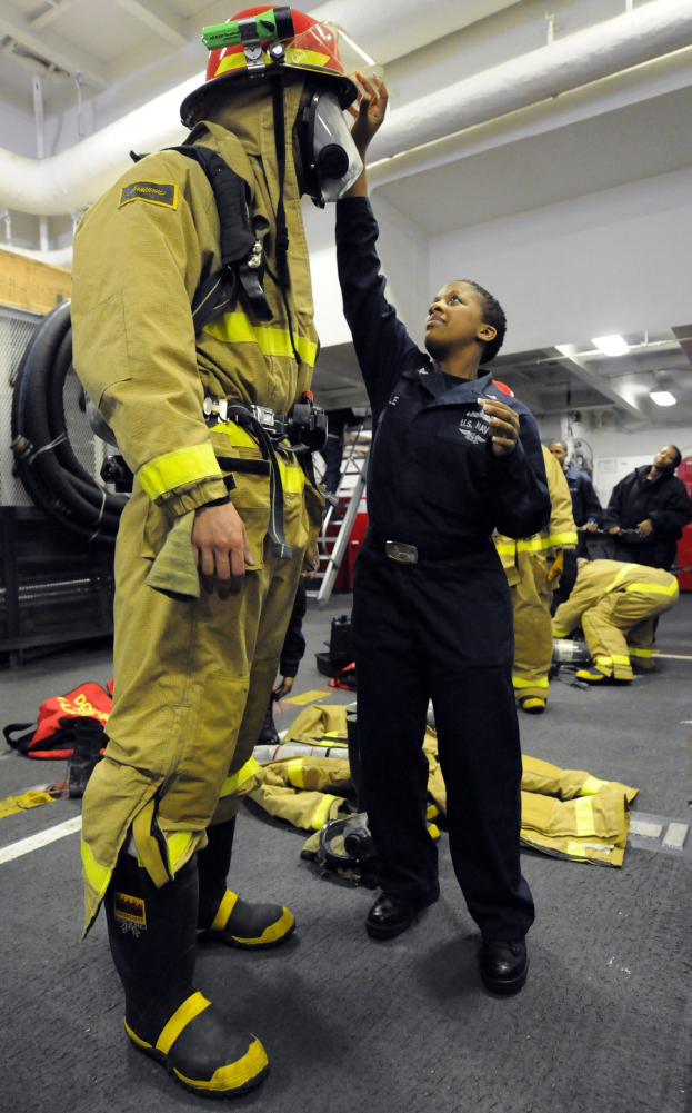 Zwei Feuerwehrleute, einer in einem schwarzen Kleid und Helm und ein anderer in gelb-schwarzer Ausrüstung, stehen neben einer Leiter und Rohren, mit anderen im Hintergrund.