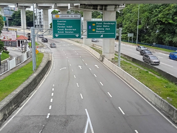 Autobahn mit fahrenden Autos, grasigem Umfeld, einem Zaun, Strommasten, Holzplanken, Bäumen, fernen Gebäuden, einer Brücke und einem sichtbaren Verkehrszeichen.