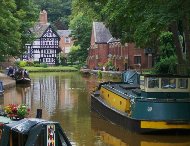 Kanal mit mehreren Booten, die an seinem Rand liegen, umgeben von grünem Gras, Pflanzen und Bäumen, mit Gebäuden im Hintergrund.