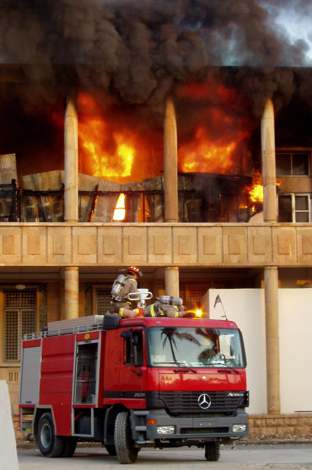 Feuerwehrl├Âschfahrzeug auf einer Stra├če vor einem brennenden Geb├Ąude mit dickem Rauch, neben einer Wand.