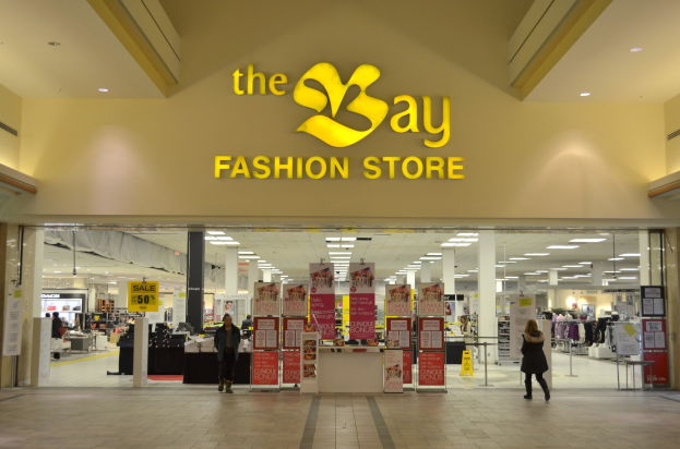 Außenansicht eines Bay Fashion Stores in einem Einkaufszentrum mit Einkäufern, Wandtexttafeln, Säulen, Deckenleuchten und Hintergrundauslagen.