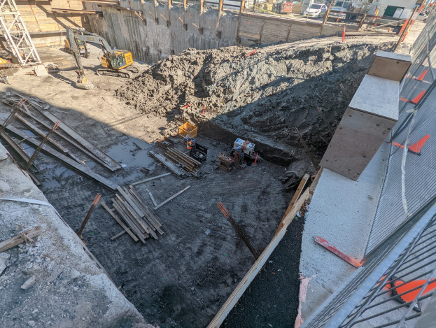 Baustelle mit einem großen Haufen Erde, umgeben von Kränen, Holzstöcken und anderem Equipment, begrenzt von einem Maschendrahtzaun, mit Gebäuden, Fahrzeugen, Pfählen und Bäumen im Hintergrund unter dem Himmel.