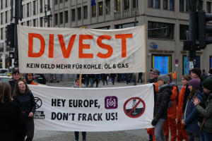 Gruppe von Menschen mit einem Banner "Divest Hey Europe, Don't Frack Us" auf einer Straße mit Gebäuden, Straßeninfrastruktur, Fahrzeugen, Bäumen und einem klaren Himmel im Hintergrund.